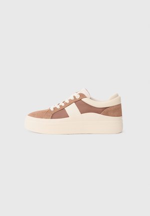 Sneaker in suede marrone con accenti crema, punta rotonda, tomaia testurizzata, lacci piatti e una suola piatta spessa di un beige chiaro.