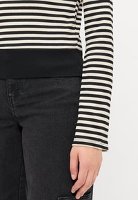 Stripet langermet topp med svarte og kremfargede horisontale striper, tettsittende ribbet tekstur, og solid svart midjebånd. Synlige svarte jeans.