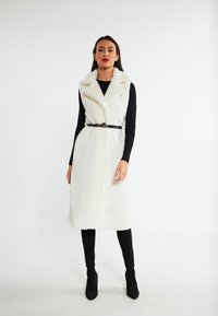 Witte nepbont mouwloze vest met een sjawlboord, strakke taille door een zwarte riem, gecombineerd met zwarte slim-fit broeken en enkellaarsjes.