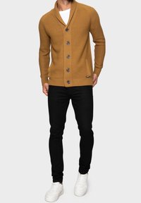 Musztardowy kardigan z dzianiny z kołnierzem szalowym, sześcioma ciemnymi guzikami i kieszenią z przodu, zestawiony z czarnymi obcisłymi dżinsami i białymi tenisówkami.