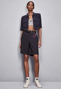 Giacca blu navy tagliata con una canotta grigia, cintura nera con un moschettone rosa e pantaloni corti neri fino al ginocchio. Sneakers alte bianche.