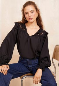 Blusa nera con colletto arricciato e dettagli a taglio, maniche lunghe con polsini elastici, abbinata a jeans blu a vita alta.
