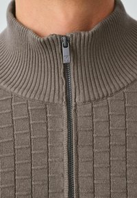 Brązowy sweter z dzianiny z wysokim prążkowanym kołnierzem i zamkiem błyskawicznym z przodu. Posiada teksturowany wzór w kratkę na korpusie oraz gładkie detale zamka.