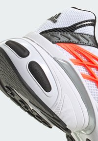 Pantofii sport albi au un superior din plasă texturată, linii de accent negre și portocalii și o talpă căptușită cu un design de profil cu model.