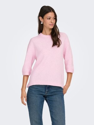 Femme aux longs cheveux bruns porte un pull rose clair à manches trois-quarts et un jean bleu, debout avec une main dans la poche.