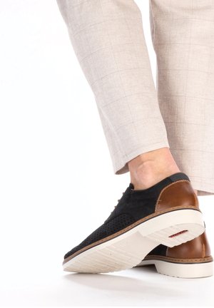 Chaussures en suede noir et cuir brun avec un design perforé, semelle en caoutchouc blanche et bout rond. Associées avec un pantalon à carreaux légers.