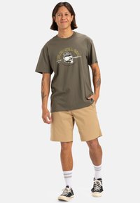 Camiseta verde oliva con estampado gráfico y logo, combinada con pantalones cortos beiges. El modelo lleva zapatos negros, calcetines a rayas blancas y tiene un ajuste holgado.