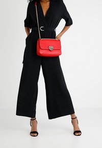 Tuta nera con gambe ampie, cintringhiata in vita. Borsa in pelle rossa con chiusura a forma di cuore, tracolla in oro e sandaletti neri open-toe.