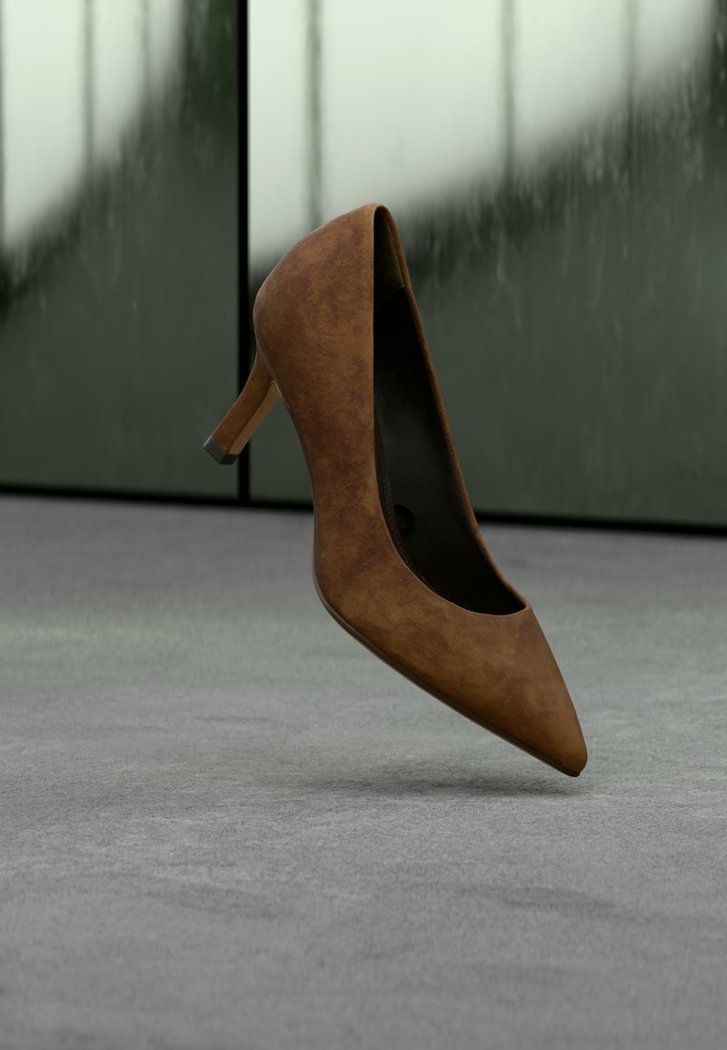 Καφέ σουέντ ψηλοτάκουνη γόβα με μυτερή μύτη, ψηλό τακούνι και απαλή υφή, παρουσιάζεται πάνω σε γκρίζα επιφάνεια με θολό φόντο.