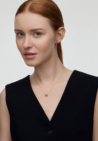 Złota naszyjnik z wisiorkiem w kształcie węzła, noszony z bezrękawnikową czarną bluzką. Modelka ma proste czerwone włosy i delikatne kolczyki wkręcane.