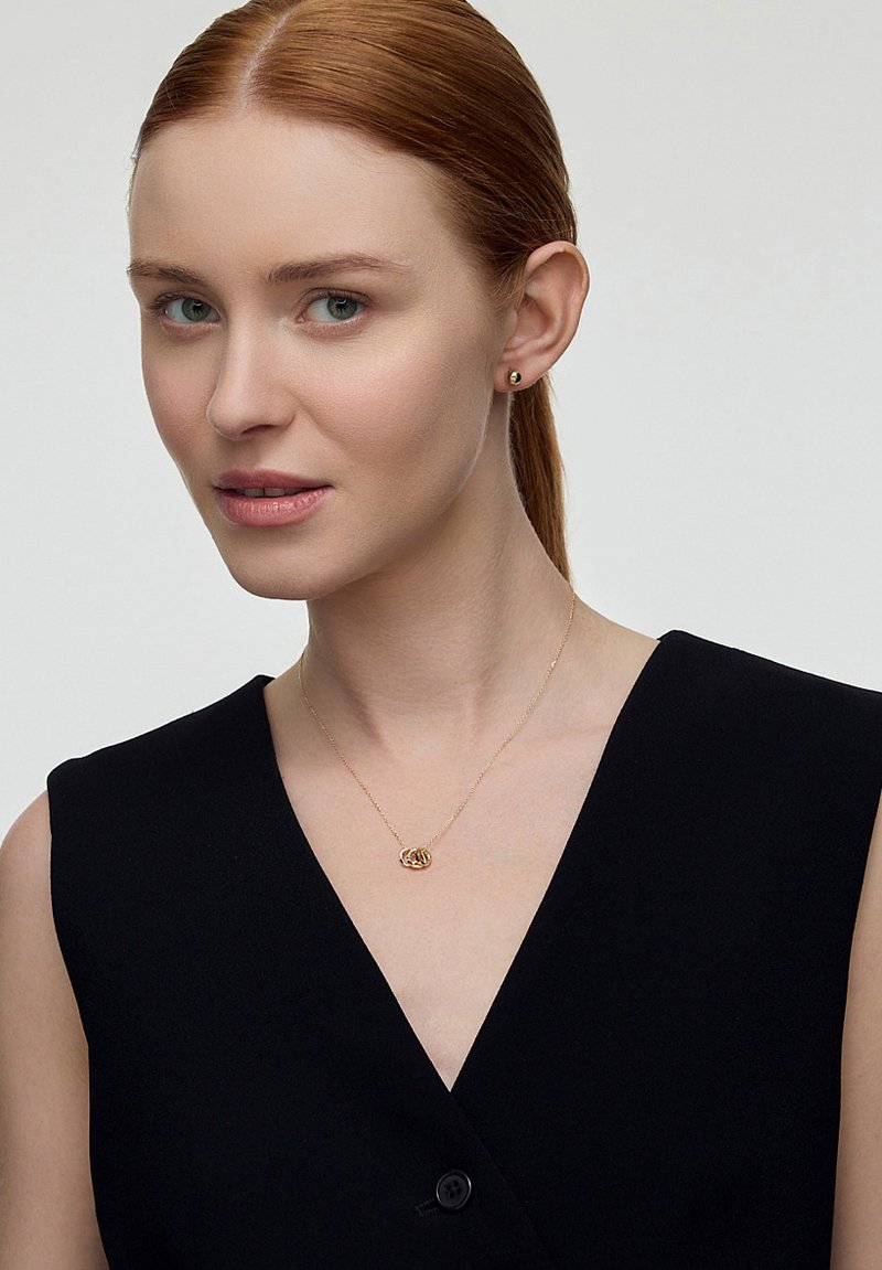 Złota naszyjnik z wisiorkiem w kształcie węzła, noszony z bezrękawnikową czarną bluzką. Modelka ma proste czerwone włosy i delikatne kolczyki wkręcane.