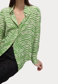 Donna che indossa una camicia a righe zebra verdi e bianche con pantaloni neri, seduta con una mano appoggiata accanto a lei.