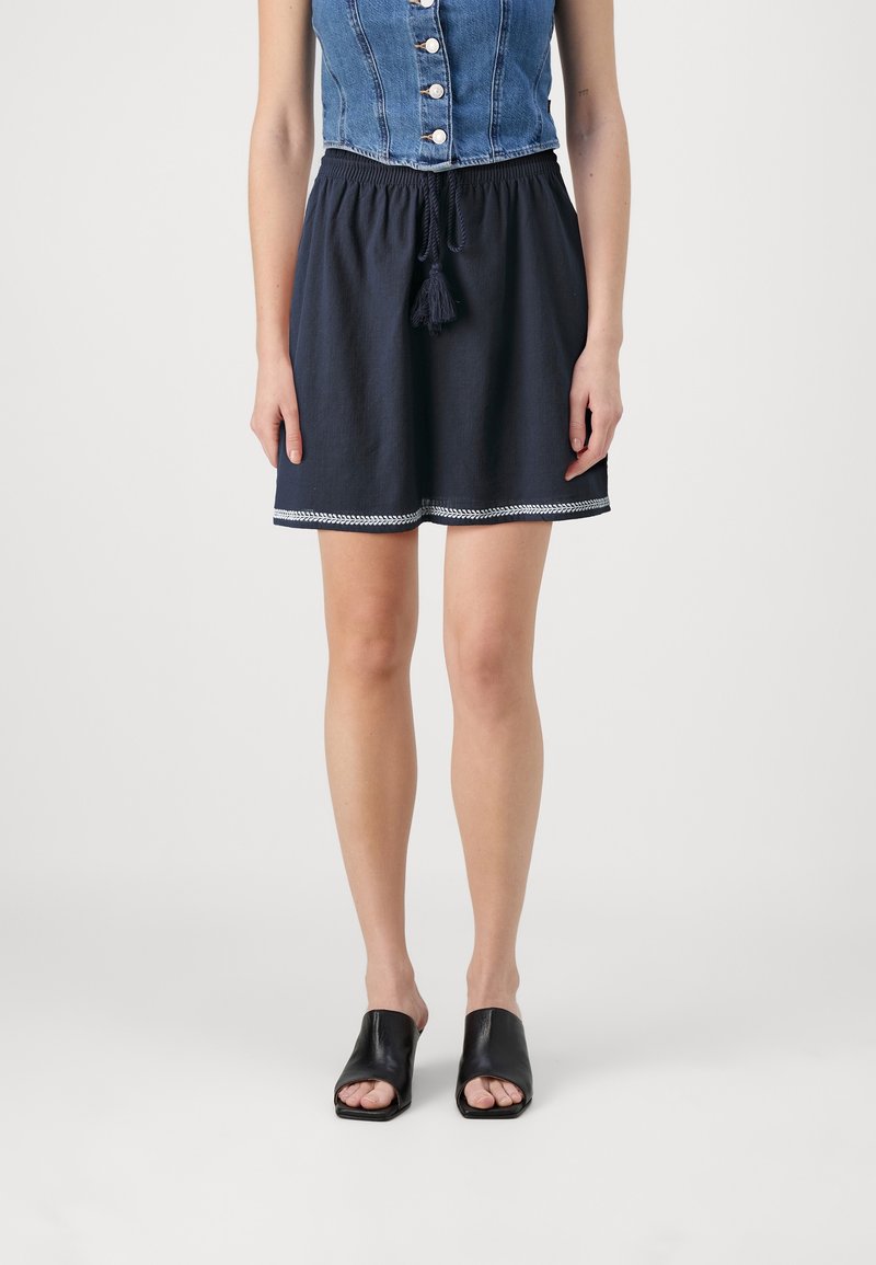 Femme portant une jupe bleu marine foncé avec des bordures blanches et un cordon à pompons, associée à un gilet en jean boutonné et des sandales mules noires à bouts ouverts.