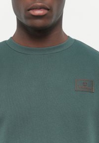Groene trui gemaakt van ribbelstof, met een ronde hals en een leren Timberland logo patch op de borst.