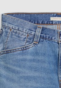 Jeans de denim de color azul claro con una cinturilla ancha, costuras naranjas y un bolsillo frontal. La tela tiene un aspecto texturizado clásico.