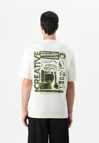 T-shirt blanc en coton à manches courtes, featuring une impression graphique en noir et vert au dos, incluant du texte et des designs abstraits.
