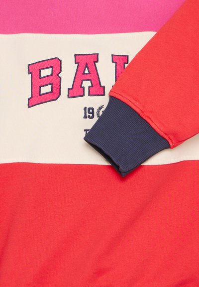 Sarkana un rozā krāsu bloka treniņjakas ar krēmveida svītru, kuras priekšpusē ir izšuvums "BALI" tumši zilā krāsā un ribota tumši zila piedurkne.