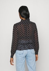 Sort gennemsigtig bluse med små blomstermønstre, samlet talje og lange ærmer. Glat tekstur og let flydende design, parret med lyseblå jeans.