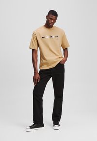 Beige, rövid ujjú póló, amely négy napszemüveges grafikus arcot ábrázol. Fekete nadrággal és fekete, fehér talpú tornacipővel párosítva.