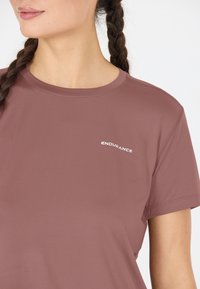 Ung kvinna med flätat hår klädd i en mauve kortärmad träningsskjorta med "ENDURANCE" tryckt på bröstet.
