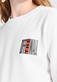 Vit sweatshirt med en strukturerad "OFF THE WALL"-logotyp i svart och röd på vänster sida. Klassisk rundhalsadesign med en avslappnad passform.