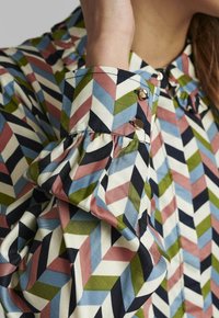 Chemise colorée présentant un motif en zigzag vert, bleu, rose et noir. Poignets volantés et accents de boutons dorés. Texture en tissu de coton.