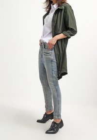 Světle olivově zelená lehká bunda, bílá košile pod ní, distressed světle modré džíny a černé boty s přezkami, ukazující ležérní styl.
