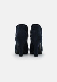 Par de botas de tacón alto de ante azul oscuro, con cremallera trasera, vistas desde el talón sobre un fondo blanco.