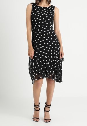 Vrouw met een mouwloze zwarte jurk met witte stippen, asymmetrische zoom en zwarte bandjes hoge hakken sandalen, staand tegen een witte achtergrond.