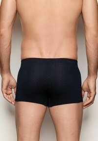 Modelo masculino visto desde atrás, vistiendo bóxer ajustados negros con pequeños lunares rojos sobre un fondo neutro.