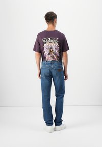 Camiseta gráfica morada con un estampado colorido en la parte posterior, combinada con jeans de mezclilla azules y zapatillas blancas, vista desde atrás.