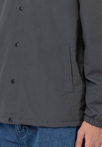 Grå knapp-up-jacka med en slät textur, svarta knappar och en sidficka. Ärmarna är elastiska och fållen är något längre.