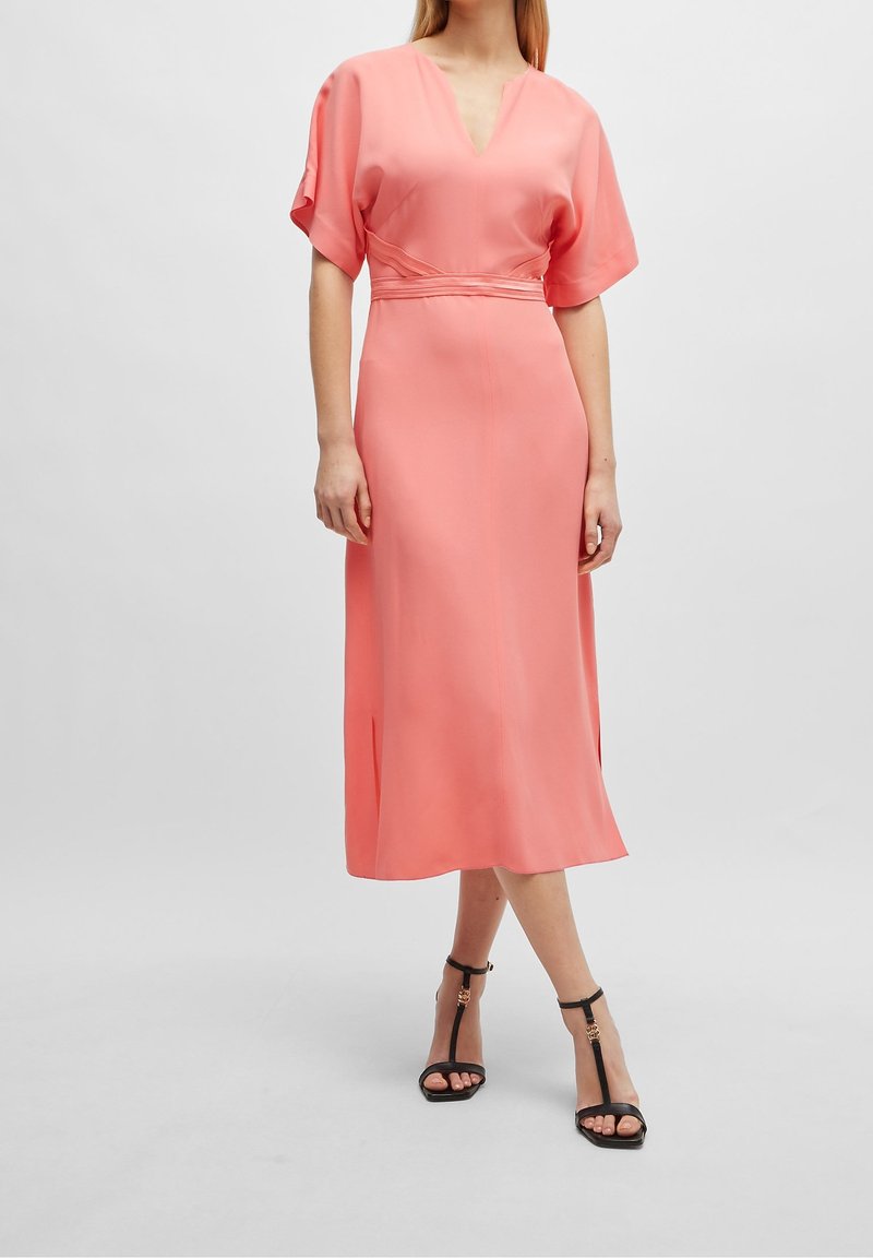 Femme portant une robe midi corail rose avec des manches aux coudes et des sandales noires à talons hauts, debout sur un fond uni.