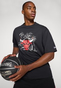 Svart bomulls t-skjorte med et rødt okse-motiv og palmer. Modellen holder en Spalding basketball, med løs passform og korte ermer.