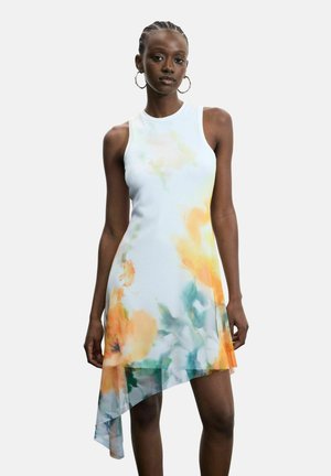 Vrouw draagt een mouwloze witte jurk met oranje en groene bloemmotieven en een asymmetrische zoom, staand tegen een effen achtergrond.