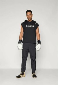 Fekete ujjatlan kapucnis pulcsi "BOXING" felirattal, fekete nadrággal és fekete-beige bokszcipőkkel párosítva. Fehér bokszkesztyűt visel.