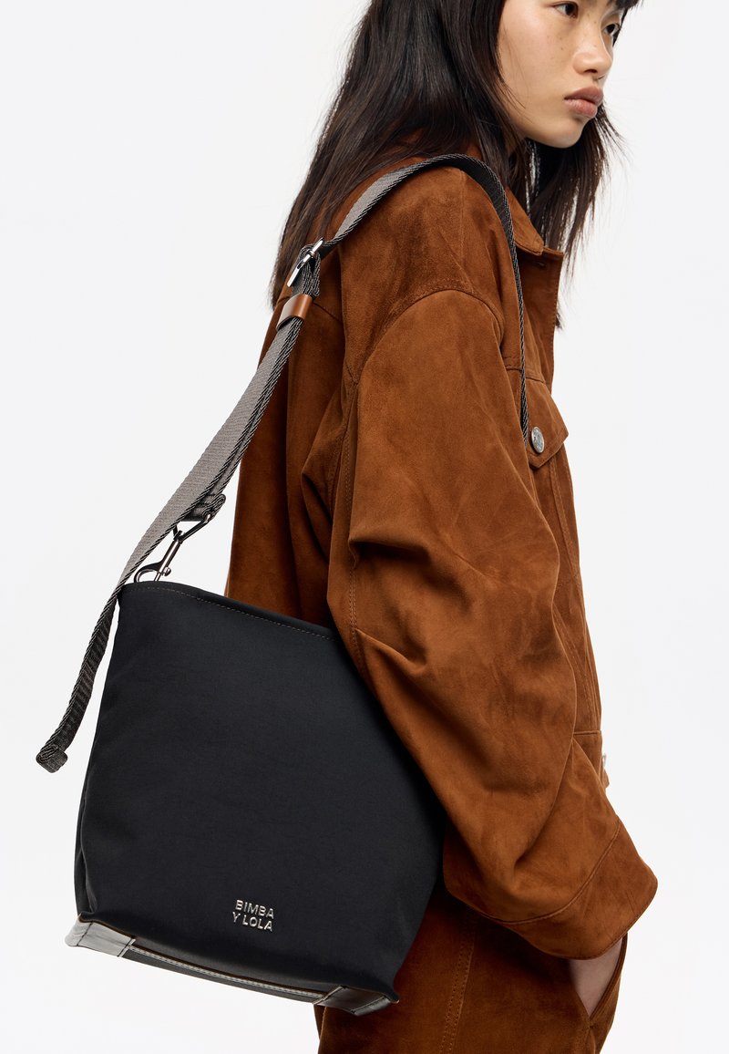 Bolsa tipo cubo de tejido negro con un diseño elegante, que presenta una correa de hombro texturizada y un detalle del logo en la parte inferior.