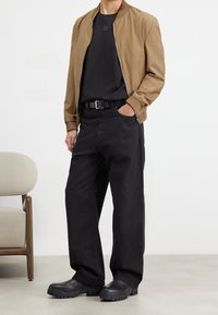 Uomo che indossa una giacca zip-up beige sopra una maglietta nera, pantaloni neri a gamba larga con cintura e stivali neri con suola spessa, in piedi accanto a una sedia beige.