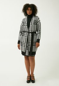 Cardigan a righe bianche e nere con cintura a nastro, caratterizzato da design geometrici e maniche lunghe, abbinato a un dolcevita nero e tacchi.