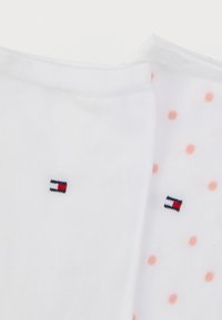 Δύο ζεύγη λευκών κάλτσες αστραγάλου, ένα απλό και ένα με ροζ πουά, κάθε ένα με ένα μικρό ορθογώνιο λογότυπο κόκκινου, λευκού και μπλε χρώματος.