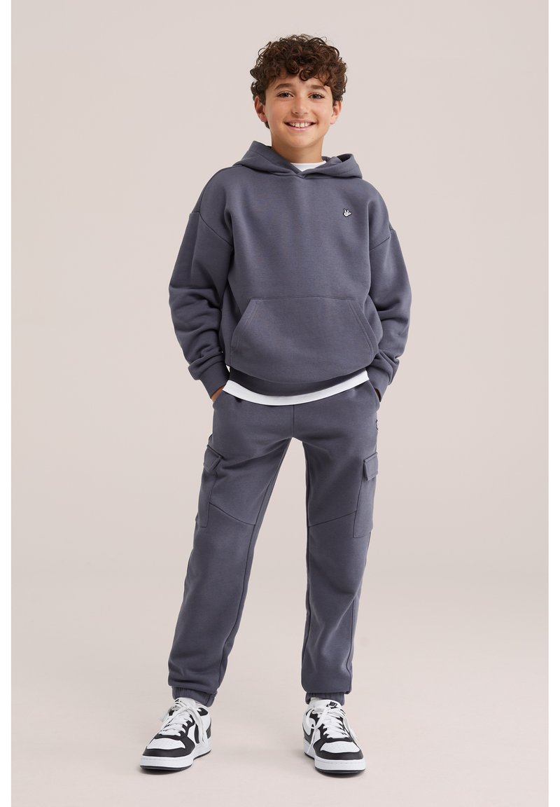 Garçon debout les mains dans les poches, portant un sweat à capuche gris foncé, un pantalon cargo assorti, un t-shirt blanc et des baskets noir et blanc, souriant.
