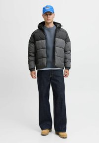 Grau gefütterte Jacke mit schwarzer Kapuze, kombiniert mit einem blauen Pullover, dunklen weiten Jeans und hellbraunen Stiefeln. Eine blaue Mütze rundet das Outfit ab.