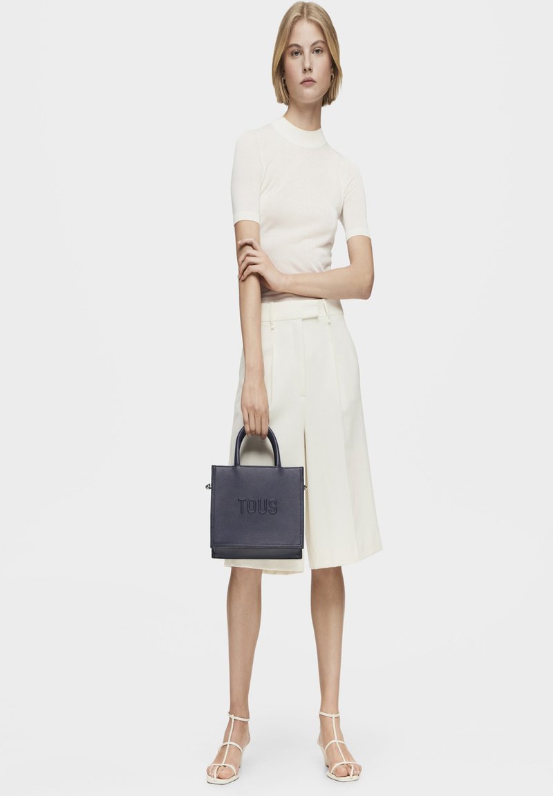 Bolso tote estructurado en piel de color marino, con el logotipo "TOUS" en relieve. El modelo lleva un top ajustado de color crema y culottes de crema clara, combinados con sandalias con tiras.