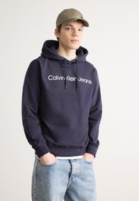 Sötétkék pamut kapucnis pulóver elöl kenguruzsebel, állítható húzózsinórok és fehér "Calvin Klein Jeans" logó. Világoskék farmernadrággal párosítva.