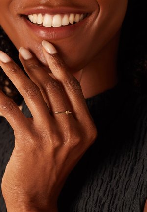 Gouden ring met een klein hartdetail op een slanke band, gedragen op een hand met amandelvormige nagels tegen een gestructureerde donkere achtergrond.