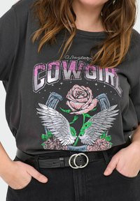 Harmaan puuvillaisen T-paidan etuosassa on graafinen design, jossa lukee "COWGIRL," sekä vaaleanpunainen ruusu, siivet ja kukka-akkentit. Yhdistetty mustan vyön kanssa.