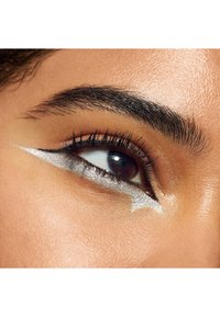 Gros plan d’un œil marron avec des cils épais et des sourcils foncés, avec un eye-liner ailé argenté qui s’étend des paupières supérieures et inférieures.