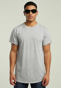 Jasnoszara koszulka z krótkim rękawem wykonana z miękkiej bawełny, z okrągłym dekoltem i małym logo na piersi, zestawiona z ciemnymi okularami przeciwsłonecznymi.