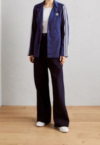 Tmavě modrý blejzr se dvěma knoflíky a bílými pruhy na rukávech, doplněný širokými tmavými kalhotami a bílými teniskami.