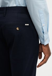 Primer plano de una persona que lleva pantalones de color azul marino oscuro con un bolsillo trasero abotonado y una camisa de manga larga azul claro.